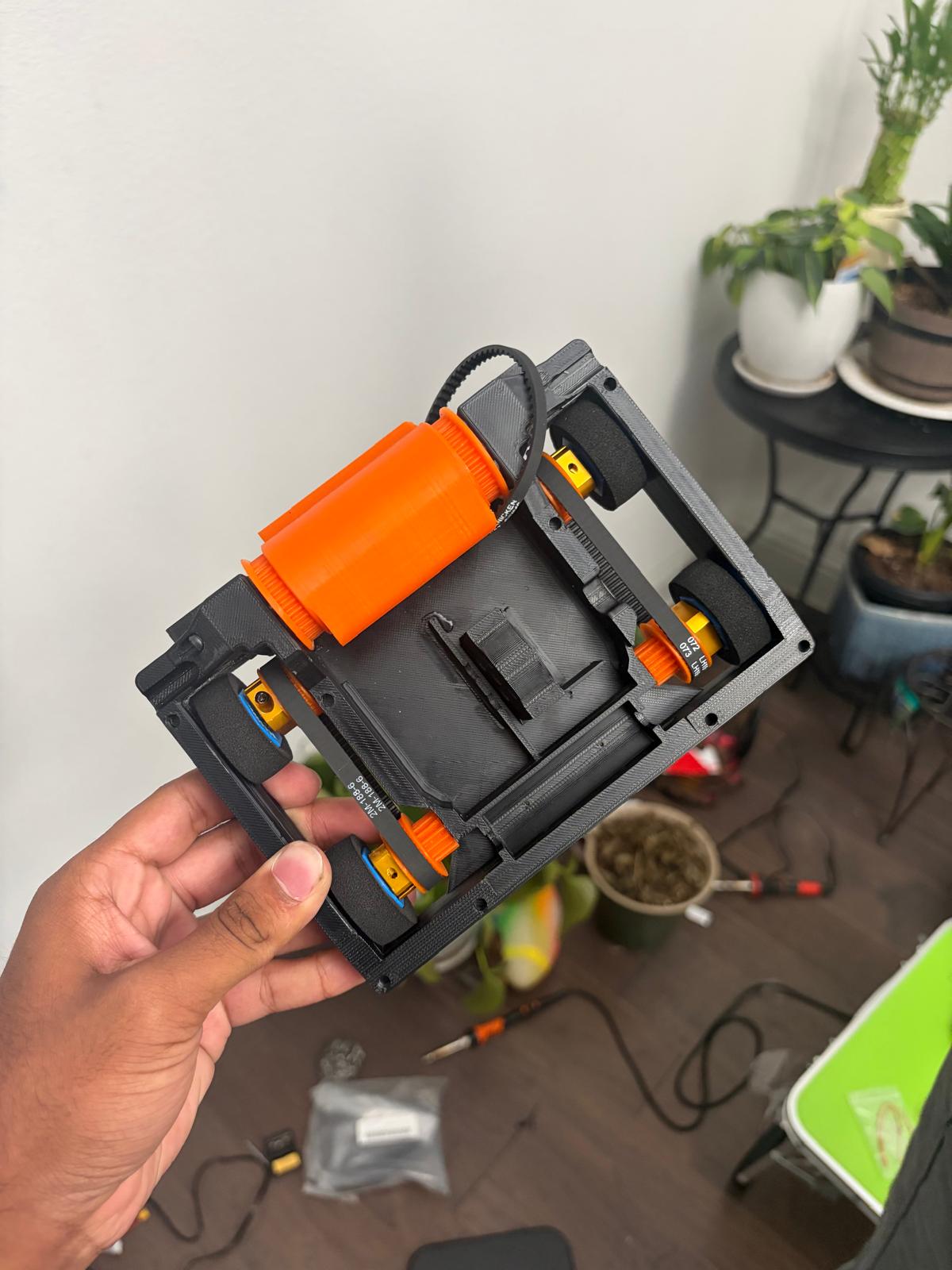 the printed chassis held in hand, top down: orange drum, belts, four foam wheels on printed pulleys, wedge